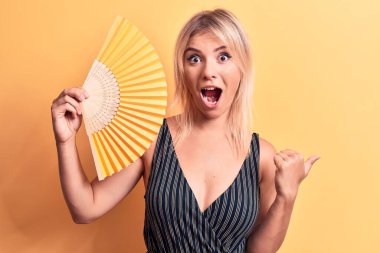 Sarışın, güzel bir kadın el fanı kullanıyor. Sarı arka planda dikiliyor. Başparmağıyla yan tarafa bakıyor.