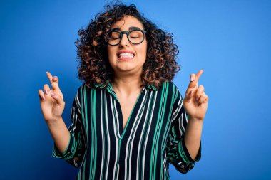 Güzel kıvırcık Arap kadın mavi arka plan üzerinde çizgili gömlek ve gözlük takıyor umut ve gözler kapalı gülümseyerek el işareti yapıyor. Şans ve batıl inanç..
