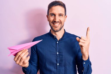 Pembe kağıt uçak tutan genç yakışıklı adam izole arka planda duruyor ve mutlu bir şekilde gülümsüyor.