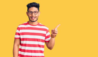 Yakışıklı Latin Amerikalı gündelik kıyafetler ve gözlük takan, yüzünde kocaman bir gülümseme olan, kameraya el ile işaret eden genç adam.. 