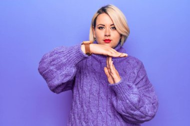 Güzel sarışın ve cüsseli bir kadın mor arka planda balıkçı yaka kazak giyiyor. Elleriyle jest yapıyor, hayal kırıklığına uğramış ve ciddi bir yüz ifadesi takınıyor.