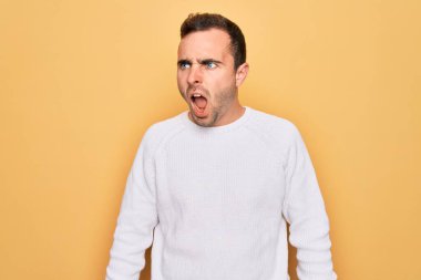 Mavi gözlü, sıradan bir süveter giyen genç bir adam sarı arka planda sinirli ve öfkeli bir şekilde bağırıyor. Öfkeyle bağırıyor. Öfke ve agresif kavram.