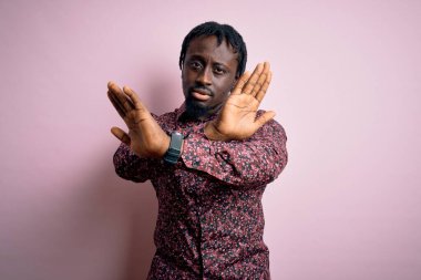 Genç, yakışıklı, Afro-Amerikalı bir adam pembe arka planda dikiliyor. Reddedilmiş yüz ifadesi, kollar ve avuçlar üzerinde negatif işaretler, kızgın yüz ifadesi.