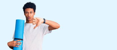 Genç Afrikalı Amerikalı adam kızgın suratlı yoga minderi tutuyor, negatif işaret baş parmakları aşağıda, reddedilme konsepti 