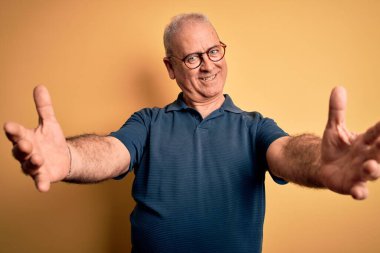 Sarı arka planda sıradan polo ve gözlük takan orta yaşlı yakışıklı adam kucaklaşmak için kollarını açıp gülümseyen kameraya bakıyor. Mutluluğu kucaklayan neşeli bir ifade..