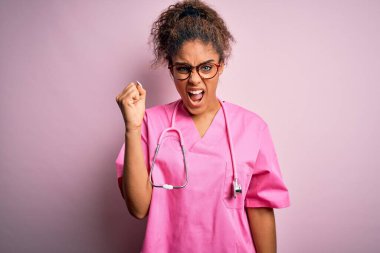 Afro-Amerikalı hemşire kız, tıbbi üniforma ve steteskopla pembe arka plan üzerinde kızgın ve kızgın. Öfkeyle bağırırken yumruğunu kaldırıyor. Öfke ve agresif kavram.