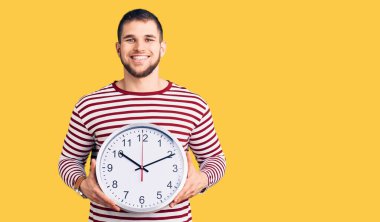 Büyük saati tutan yakışıklı genç adam pozitif ve mutlu görünüyor. Duruyor ve gülümsüyor. Kendine güvenen bir gülümsemeyle dişlerini gösteriyor. 