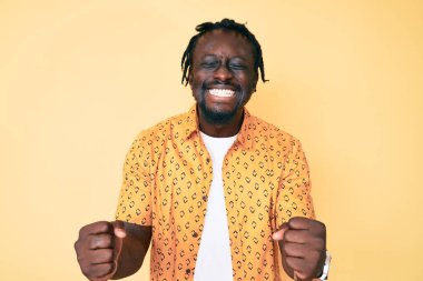 Saçları örgülü, mutlu ve heyecanlı genç Afro-Amerikalı adam kolları havada, gülümseyerek ve başarı için çığlık atarak zafer jesti yapıyor. kutlama konsepti. 