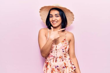 Genç, güzel, Latin bir kadın, yaz şapkası takıyor. Gülümsüyor, eli ve parmağı yana doğru uzanıyor. 