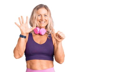 Orta yaşlı sarışın kadın spor kıyafetleri giyiyor ve kulaklık takıyor. Altıncı parmaklarını gösterip işaret ederken kendinden emin ve mutlu gülümsüyor.. 