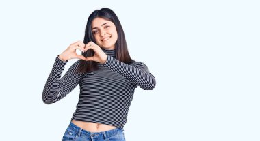 Çizgili tişört giyen genç ve güzel bir kız. Sevgiyle gülümsüyor. Elleriyle kalp sembolü ve şekli gösteriyor. romantik kavram. 