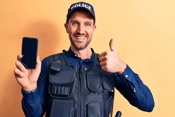 Handsome policeman wearing uniform and bulletprof holding smartphone showing screen smiling happy and positive, thumb up doing excellent and approval sign