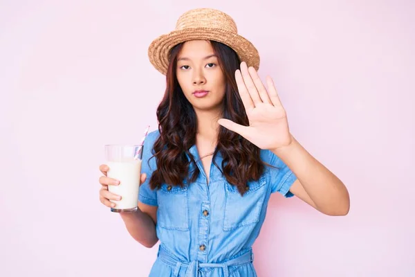 年轻美丽的中国姑娘头戴夏帽 张开手拿着一杯牛奶 做手势 严肃而自信 做手势防守 — 图库照片