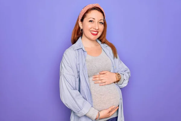 顔に幸せとクールな笑顔で孤立した紫色の背景に赤ちゃんを期待若い美しい赤毛の妊婦 運のいい人 — ストック写真