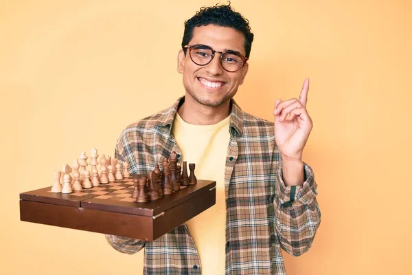 Joven Hombre Hispano Guapo Sosteniendo Tablero Ajedrez Con Gafas Sorprendidas — Foto de Stock