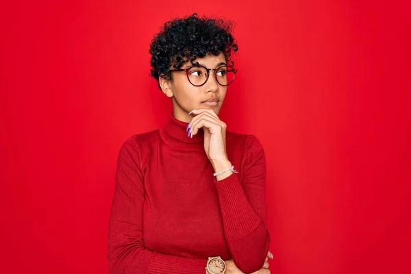 Young beautiful african american afro woman wearing turtleneck sweater and glasses with hand on chin thinking about question, pensive expression. Smiling with thoughtful face. Doubt concept.