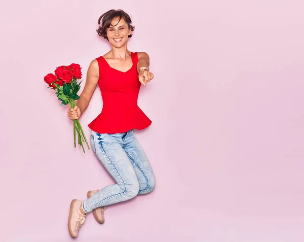 若い美しい女性のバラの幸せな笑みを浮かべて花束を保持 顔に笑顔でジャンプ上の指で指差すピンクの背景 — ストック写真