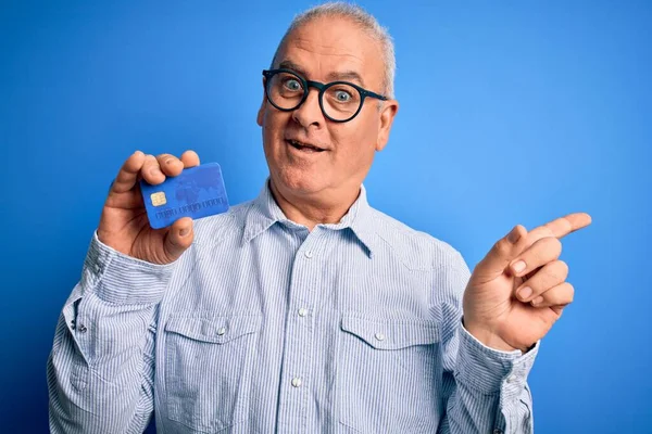 Middle age hoary man wearing glasses holding credit card as money to do payment very happy pointing with hand and finger to the side