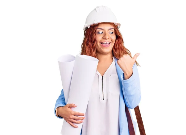 年轻的拉丁女人戴着建筑师的硬礼帽 头戴皮包 手里拿着设计图 把大拇指指向侧面 笑容满面 张开嘴 — 图库照片