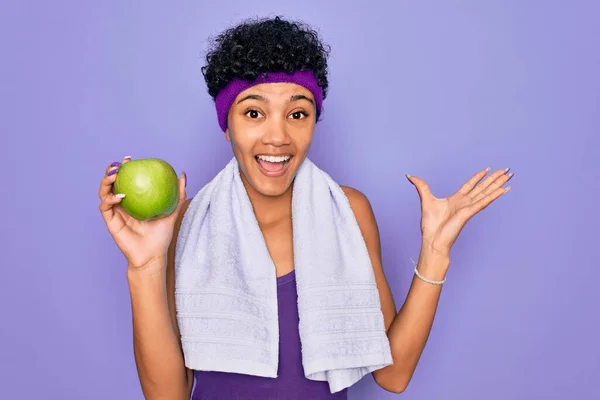 Beautiful african american afro sporty woman doing exercise wearing towel eating green apple very happy and excited, winner expression celebrating victory screaming with big smile and raised hands