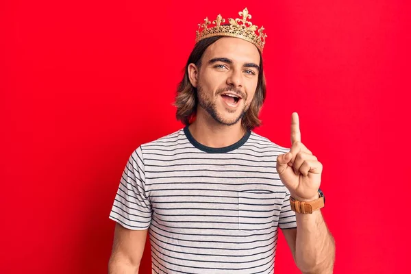 Young handsome man wearing prince crown smiling with an idea or question pointing finger up with happy face, number one