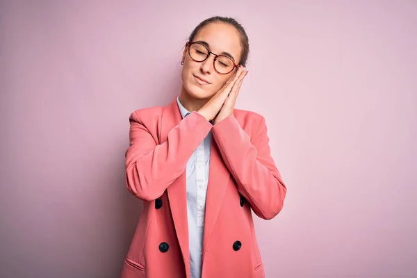 Young beautiful businesswoman wearing jacket and glasses over isolated pink background sleeping tired dreaming and posing with hands together while smiling with closed eyes.