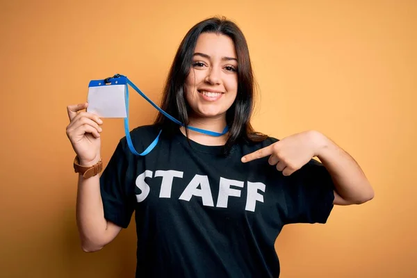 Young brunette worker woman wearing staff t-shirt as uniform showing id card with surprise face pointing finger to himself
