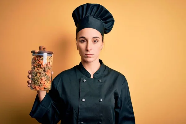 Young beautiful chef woman wearing cooker uniform and hat holding bottle with italian pasta with a confident expression on smart face thinking serious