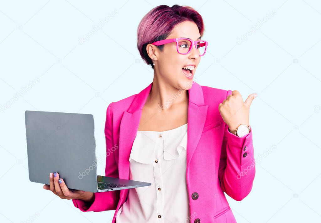 Young beautiful woman with pink hair wearing glasses holding laptop pointing thumb up to the side smiling happy with open mouth 
