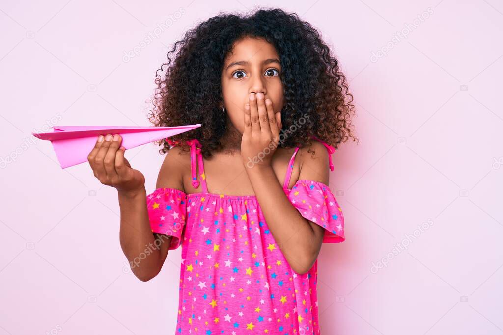 African american child with curly hair holding paper airplane covering mouth with hand, shocked and afraid for mistake. surprised expression 