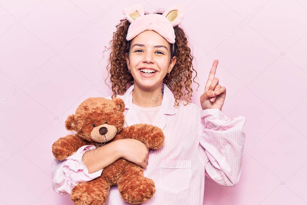 Beautiful kid girl with curly hair wearing sleep mask and pajamas holding teddy bear surprised with an idea or question pointing finger with happy face, number one 
