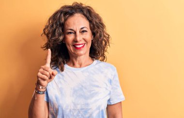 Orta yaşlı güzel bir kadın sarı arka planda dikilen günlük boyalı tişört giyiyor. Bir fikir ya da soruyla gülümsüyor ve mutlu bir yüzle işaret ediyor.