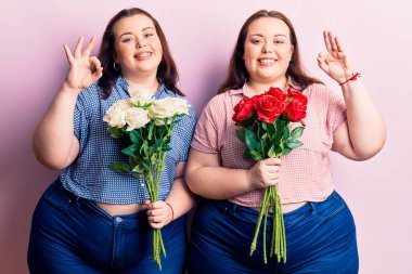 Çiçek tutan genç ve büyük beden ikizler parmaklarıyla işaret yapıyor, gülümseyerek mükemmel bir sembol oluşturuyor. 