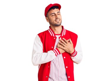 Beyzbol üniforması giyen, elleri göğsünde, gözleri kapalı gülümseyen ve yüzünde minnet ifadesi olan genç İspanyol bir adam. sağlık kavramı. 