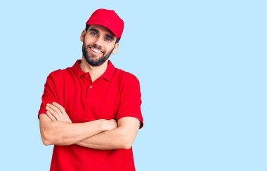 Sakallı, teslimat üniforması giyen, kollarını kavuşturup kameraya gülümseyen yakışıklı bir genç. pozitif kişi. 