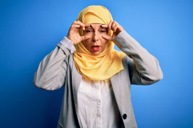 Orta yaşlı esmer iş kadını mavi arka plan üzerine geleneksel başörtüsü takıyor parmaklarıyla gözlerini açmaya çalışıyor, uykulu ve yorgun sabah yorgunluğu için.