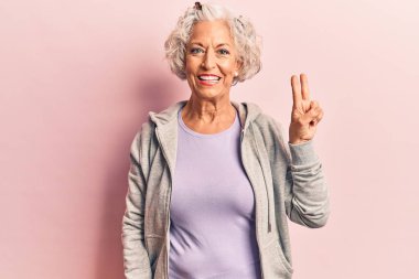 Son sınıf gri saçlı, sıradan spor giysiler giyen, kameraya gülümseyen ve zafer işareti yapan bir kadın. İki numara.. 