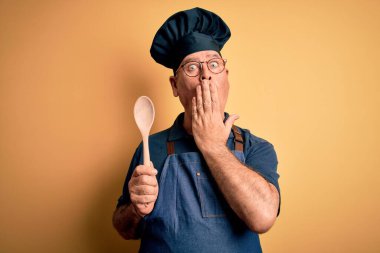Orta yaşlı tencereci önlük ve şapka takıyor sarı arka planda tahta kaşıkla ağzı kapatıyor hata yüzünden utançla dolu, korkunun ifadesi, sessizlikten korkmak, gizli konsept
