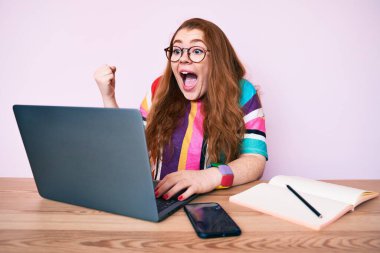 Ofiste çalışan genç kızıl saçlı kadın dizüstü bilgisayarla gururla bağırıyor zaferi kutluyor ve başarıyı kollarını kaldırarak kutluyor. 
