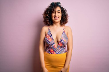 Genç, güzel bir Arap kadın, mayo ve pembe arka planda güneş gözlüğü takmış seksi bir ifadeyle kameraya bakıyor, neşeli ve mutlu bir yüzle..