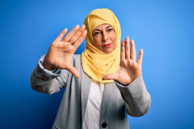 Orta yaşlı esmer iş kadını. Mavi arka plan üzerine geleneksel başörtüsü takıyor. El ve parmakları kullanarak çerçeve yapıyor.