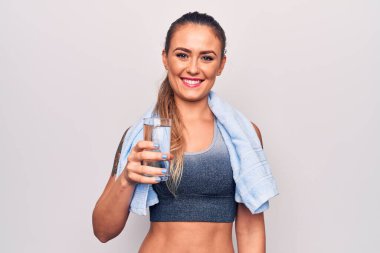 Havluyla beyaz arka planda su içen güzel sarışın kadın pozitif ve mutlu görünüyor ve kendine güvenen bir gülümsemeyle dişlerini gösteriyor.