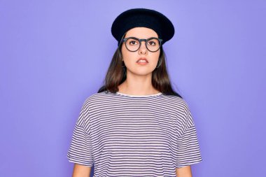 Genç, güzel, esmer, gözlüklü ve Fransız bereli bir kadın mor arka planda, yüzünde ciddi bir ifade var. Basit ve doğal bir şekilde kameraya bakıyorsun..