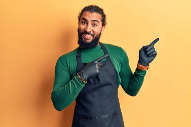 Genç Arap adam berber önlüğü takıyor gülümsüyor ve kameraya bakıyor iki eliyle ve parmaklarıyla kenarı işaret ediyor.. 