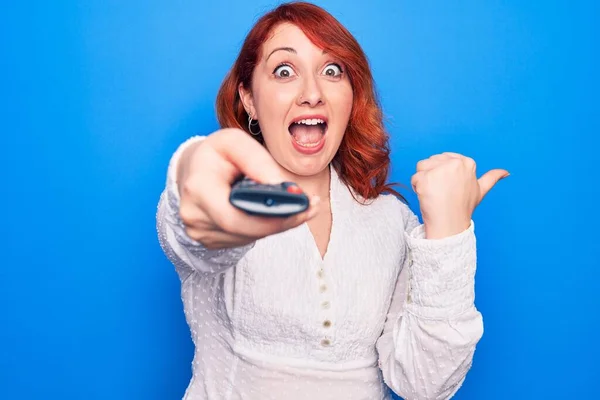 Young beautiful redhead woman changing television channel using tv remote control pointing thumb up to the side smiling happy with open mouth