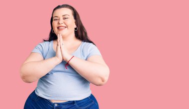 Genç ve cüsseli bir kadın gündelik giysiler giyer ellerle dua eder af diler kendinden emin bir şekilde gülümseyerek.. 