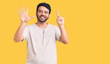 Latin asıllı genç adam gündelik giysiler giyip altı numaralı parmaklarıyla işaret ederken kendinden emin ve mutlu bir şekilde gülümsüyor.. 