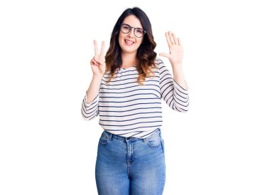 Güzel, genç, esmer bir kadın günlük kıyafetler ve gözlük takıyor. Yedi numaralı parmaklarıyla gösteriyor ve işaret ediyor. Kendinden emin ve mutlu bir şekilde gülümsüyor.. 