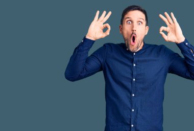 Sıradan gömlek giyen genç yakışıklı adam şaşırmış ve şaşırmış görünüyor. Tamam sembolünü parmaklarla yapıyor. çılgın ifade 