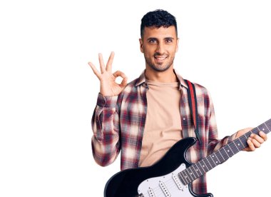 Elektro gitar çalan genç İspanyol adam parmaklarıyla imza atıyor, gülümseyerek mükemmel bir sembol oluşturuyor. 